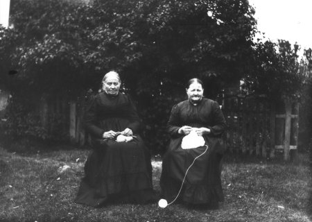 To gamle damer som sitter ute og strikker. Bildet er tatt av H.H. Lie og tilhører Maihaugens fotoarkiv.